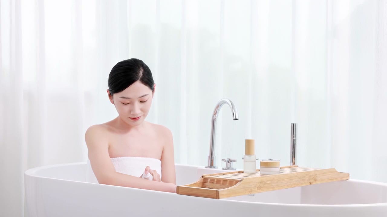 タオルに包まれた魅力的で上品な女性が手に保湿ボディローションを塗っている。バスルームのバスタブに座って首の肌に保湿クリームを塗る女性。なめらかな肌のセルフケア習慣のコンセプト。