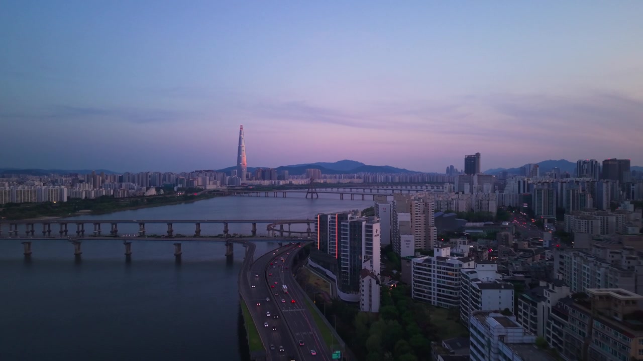 Volo aereo in avanti sopra la tortuosa superstrada Gangbyeonbuk-ro al tramonto, con i ponti del fiume Han e la luminosa Lotte World Tower sullo skyline rosa-violaceo di Seul al crepuscolo.