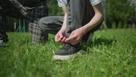 leg view of a person adjusting their shoelace while squatting on a grassy field, with a partial view of another person kneeling on one leg and a partial view of someone in red clothing