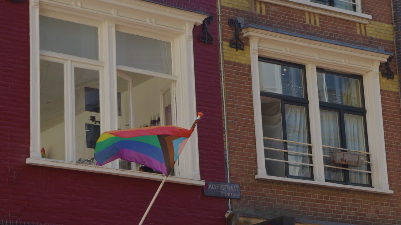 Rainbow pride flag flutters in breeze outside traditional Amsterdam building, daylight, static camera