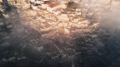 drone shot of old italian city town - matera. the city is shrouded with low handing clouds, buildings are visible through them. warm colours of the rising sun reflected against buildings