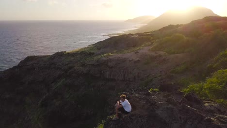 man resting on top of a cliff at sunset hiking in makapuu hawaii - aerial dolly back tilt