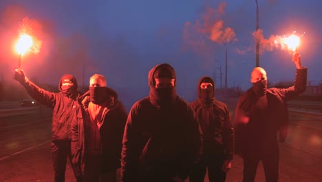 group of young men in balaclavas with red burning signal flare walking on the road under the bridge, slow motion