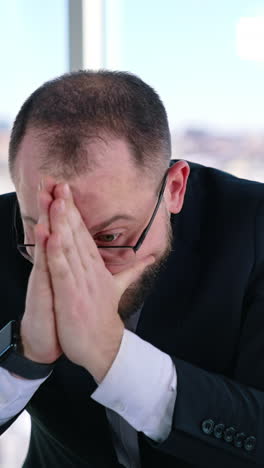 depressed businessman sits at desk. bearded middle aged man in glasses with his hands on head sitting at workplace and thinks. portrait of sad businessman in office. vertical video