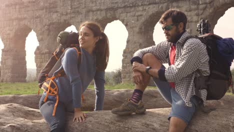 happy young couple backpackers tourists sitting on a log trunk talking and smiling in front of ancient roman aqueduct ruins in parco degli acquedotti park in rome at sunrise with guitar sleeping bag slow motion steadycam