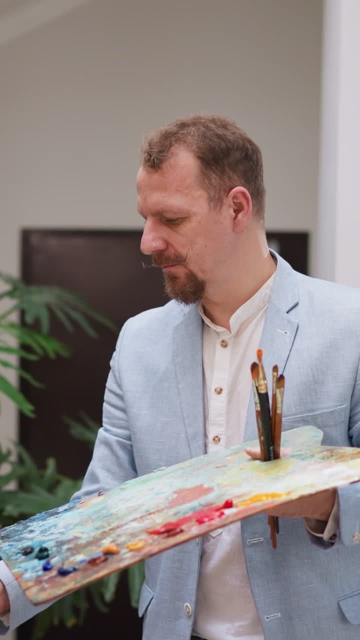 Artist with color palette works in studio. Thoughtful man in blue jacket draws applying pigment smears onto canvas on easel. Professional painter job