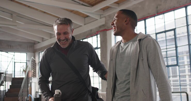 Two men entering loft, older man starting chat, gesturing with bottle and guiding pal to camera