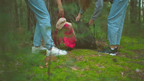 primer plano de excursionistas en vaqueros y zapatillas deportivas alcanzando sus mochilas colocadas en el musgo verde exuberante en la base del árbol en un entorno forestal tranquilo, preparándose para su viaje