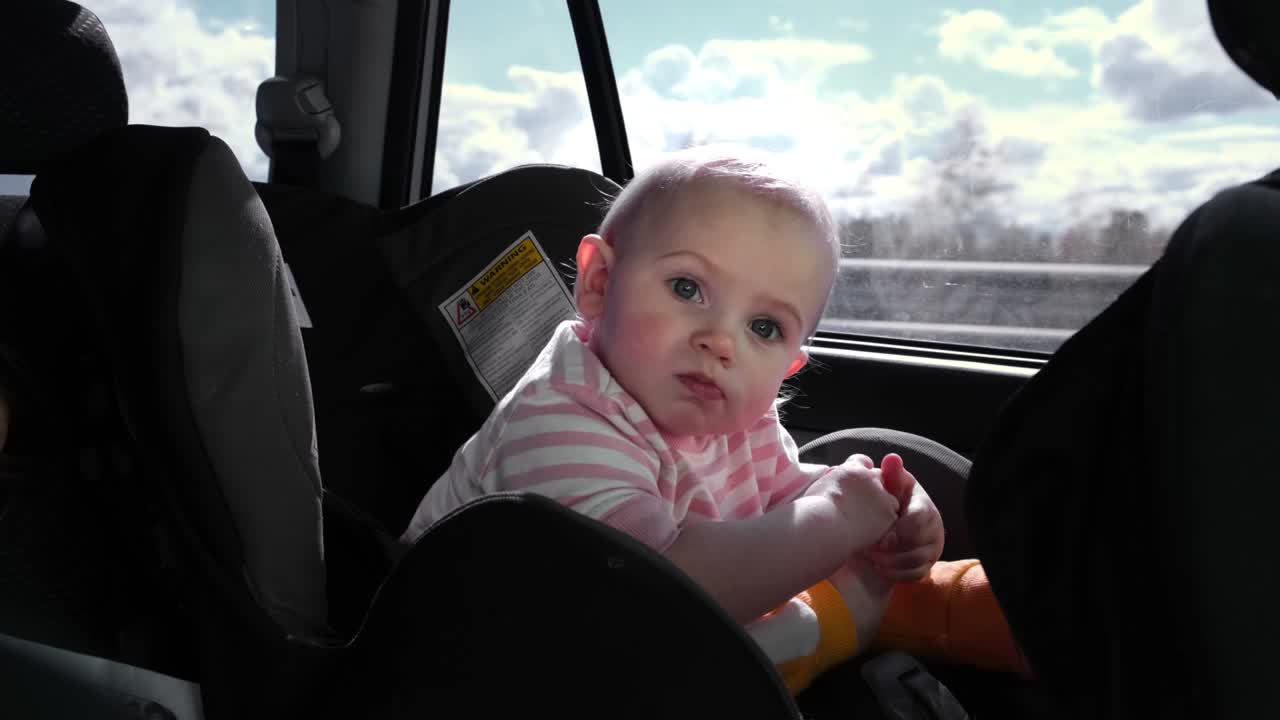 Little girl sitting in baby car seat while the car is driving. Baby travelling in a carseat in the back of vehicle. Long car trip. Little child playing, drinking. Tourism and travel, journey concept.