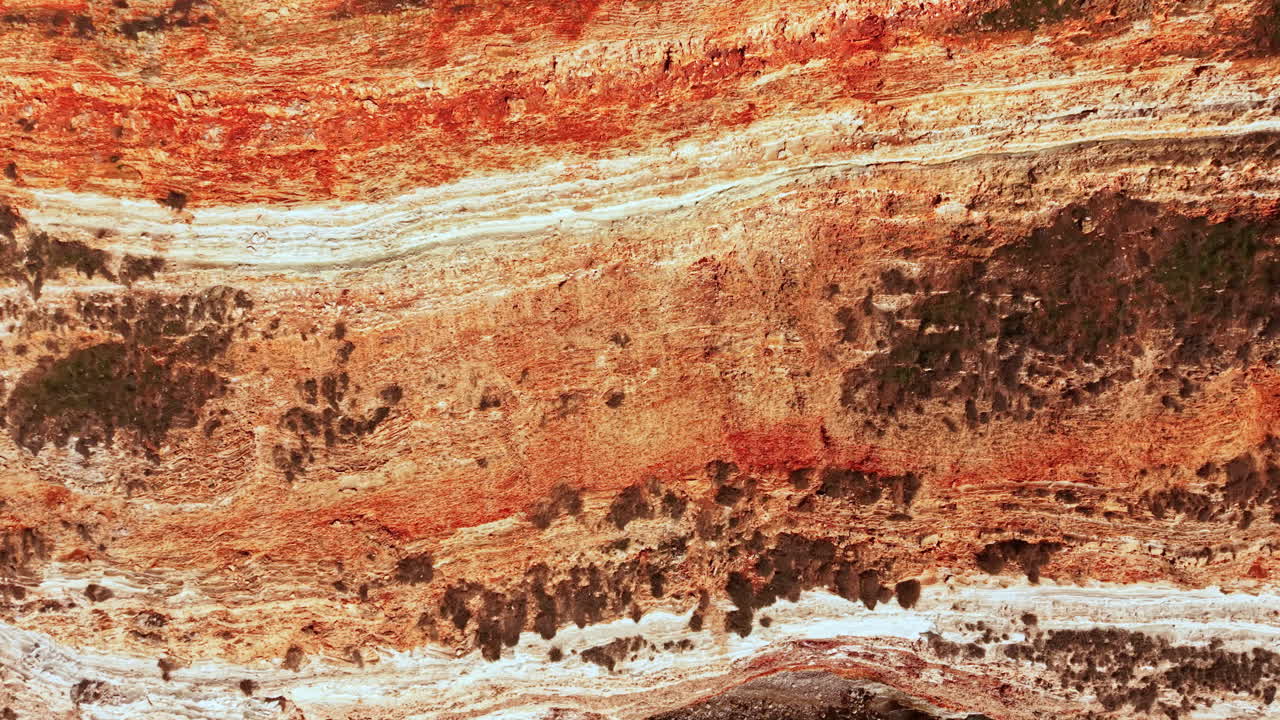 Strati di roccia e sedimenti sono visibili sulla parete di una scogliera in un ambiente naturale durante le ore diurne, mostrando colori e trame diversi derivanti da vari processi geologici