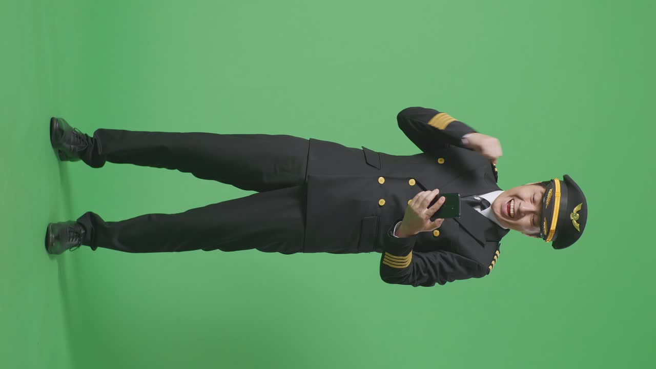 아시안 남성 조종사가 스마트폰을 쳐다보며 소리를 지르며 골을 축하하며 녹색 스크린 배경 스튜디오에 서 있습니다.