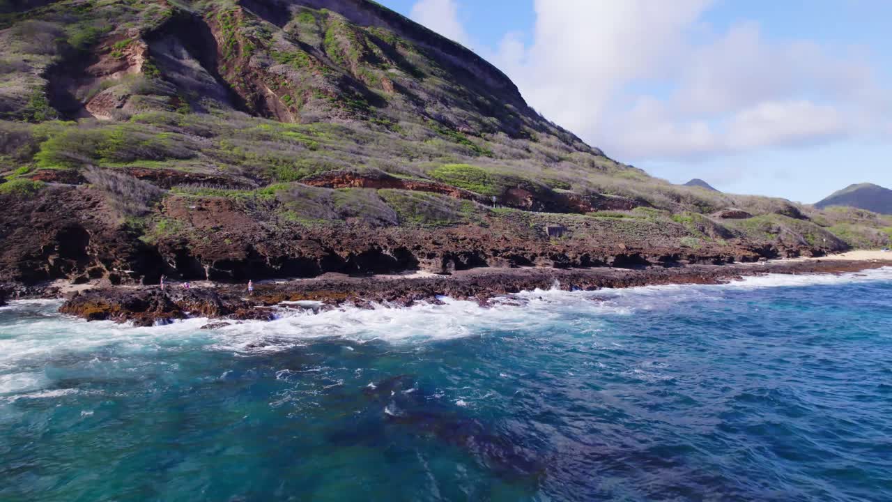 drone footage across the turquoise water of the Pacific Ocean to the coastline of Oahu in the Hawaiian Islands with white capped waves crashing onto the rocky shore of the volcanic island