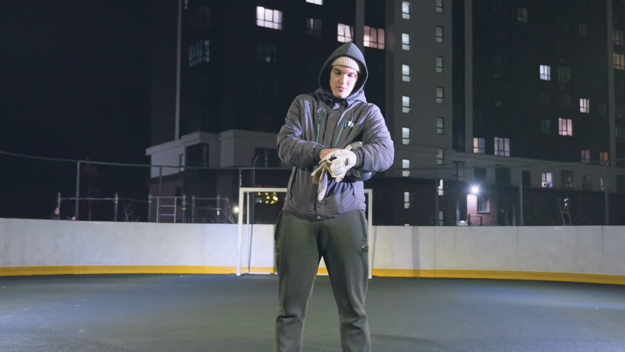 Goalkeeper prepares for action by wearing gloves on urban field during nighttime, distant background features two walkers and lit residential buildings