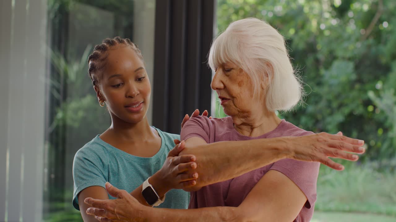 Diverse female caregiver placing hands behind senior, guiding arm stretch by glass wall smartwatch