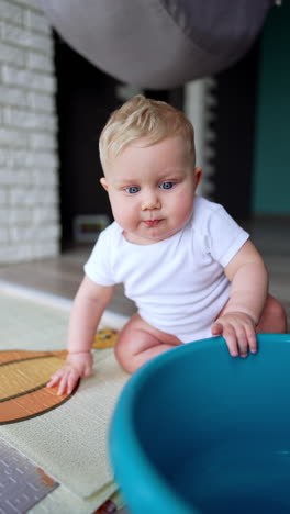 little infant kid sits on the floor covered with blue washtub. blond baby drops the wash bowl. vertical video.