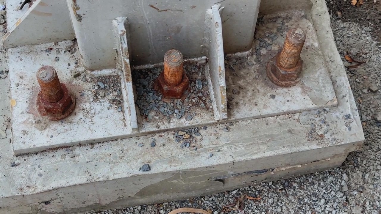 Inquadratura dinamica con telecamera orbitante di bulloni di ancoraggio in acciaio arrugginito montati su una fondazione in cemento in un cantiere industriale con elementi strutturali dettagliati e terreno ghiaioso