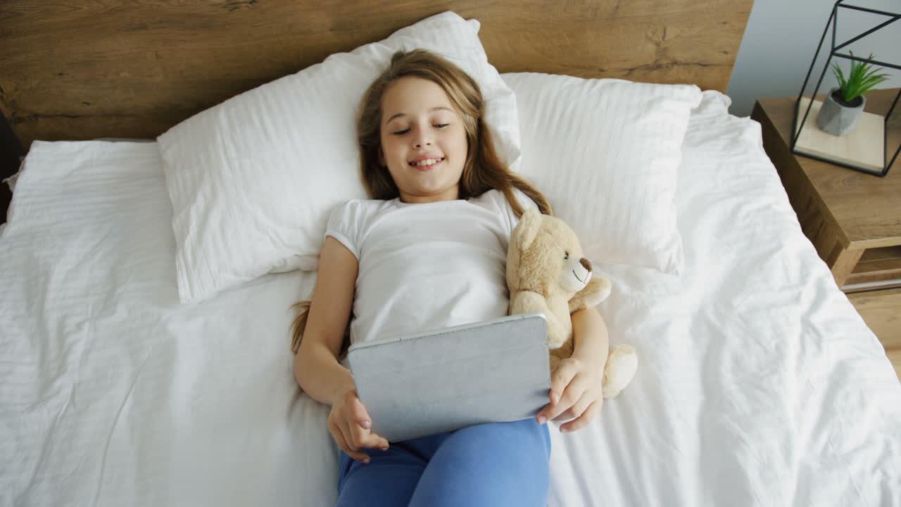 아침에 침대에 누워 웃고 태블릿에서 무언가를 보고 있는 작고 예쁜 소녀의 상위 뷰