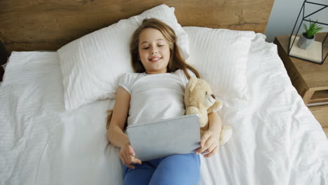 top view of a little pretty girl lying on the bed in the morning, smiling and watching something on the tablet