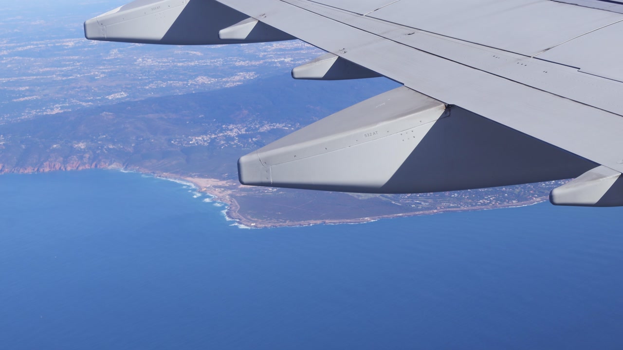 Overlooking airplane window view with wing tilt during flight over blue Atlantic ocean water showing Portugal rugged coastline land and hazy morning light from high altitude. Plane reaching Lisbon