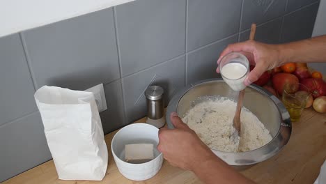 la mano del hombre mediterráneo vierte un vaso de leche sobre harina blanca dentro de un recipiente de metal.