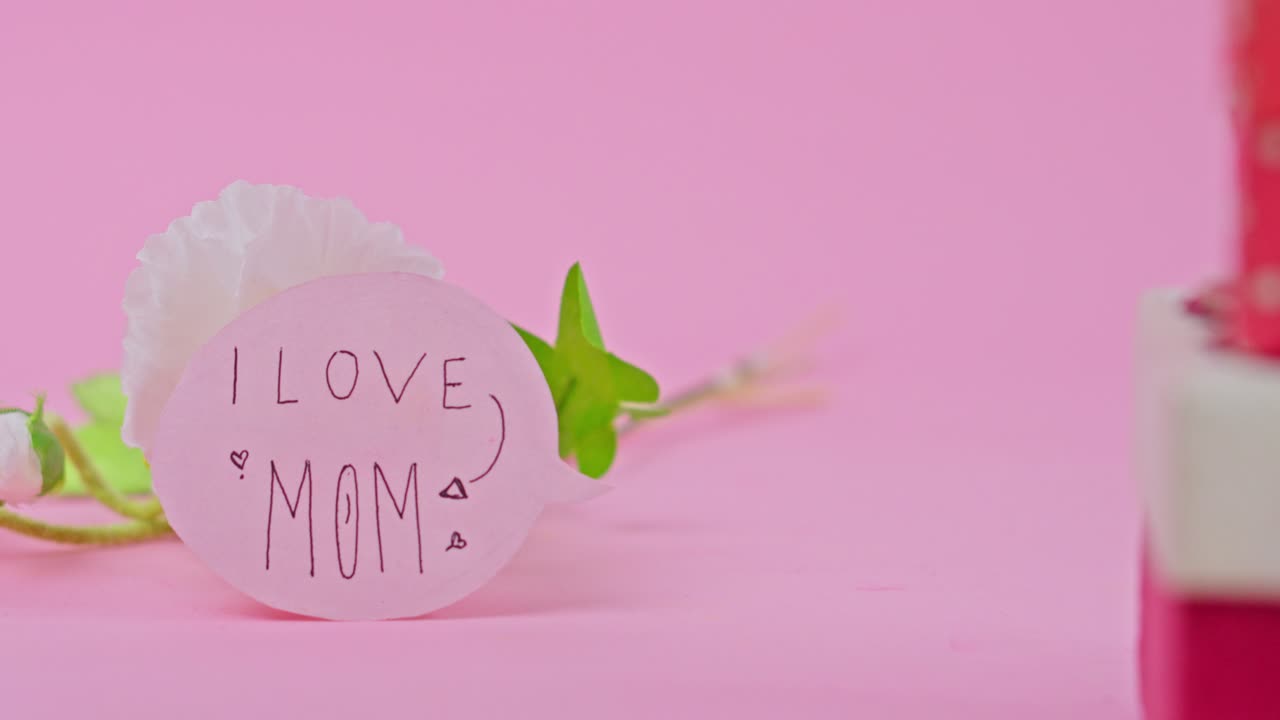 Mother's Day scene featuring handwritten "I LOVE MOM" note white carnation and gift boxes