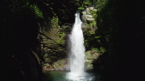 Majestic-zen-summer-scene-looking-at-Saltos-Jima-rocky-cliffside-waterfall-flowing-down-rocky-cliff-in-shadowed-forest,-Dominican-Republic,-static-low-angle