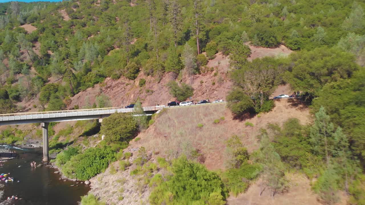 alejarse de un puente y autos a la ladera de una montaña rodeada de árboles verdes y llena de gente nadando en el río - disfrutando su fin de semana de verano