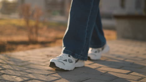 close-up shot of a person wearing jeans and white shoes, walking slowly on a paved path in a park. the steps are deliberate and careful, set against the backdrop of soft light and a blurred background