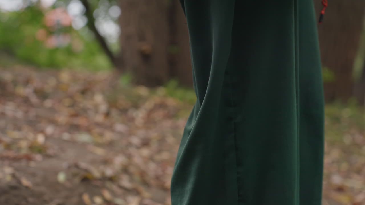 Donna bianca che percorre un sentiero nel bosco in autunno, indossa pantaloni verdi e una giacca scura, studentessa che vaga su un terreno pieno di foglie, scarpe che scricchiolano sul terreno, si ferma per sedersi su una radice muschiosa, treccia sulla nuca, profilo