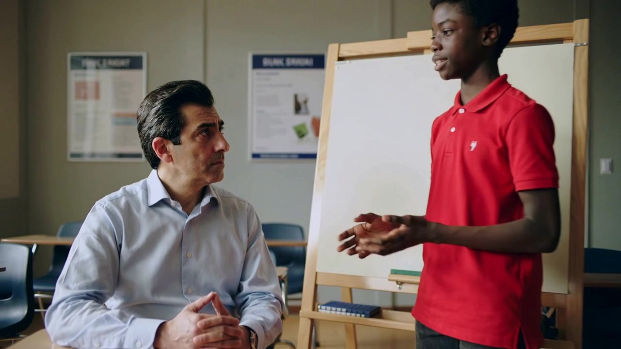 Presenting student in red polo stepping up and explaining work at easel in class, teacher listening