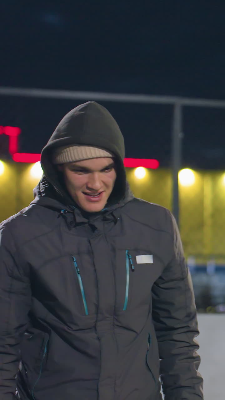 молодой человек в капюшоне улыбается с сосредоточенным выражением лица во время ночной деятельности на открытом городском поле, на заднем плане освещенный забор, яркие желтые и красные огни