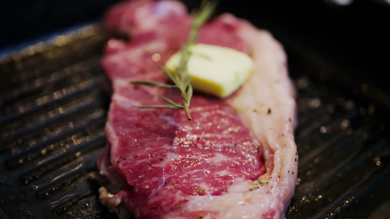 Mantequilla derritiéndose sobre un bistec sazonado cocinándose en una sartén grill caliente con hierbas de romero