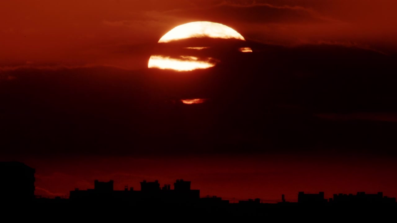 Wide shot of the sun slowly lowering behind a dense band of dark clouds, with a glowing horizon line beneath