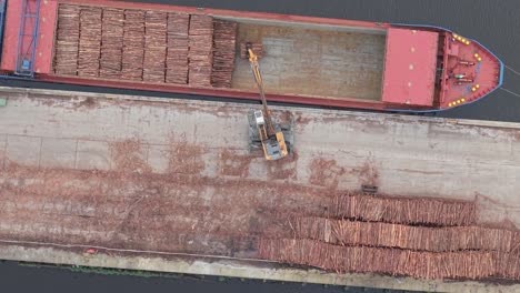 aerial view of hydraulic loader loading stacked logs into cargo ship at dockside lumber yard in port of limerick, ireland. topdown shot