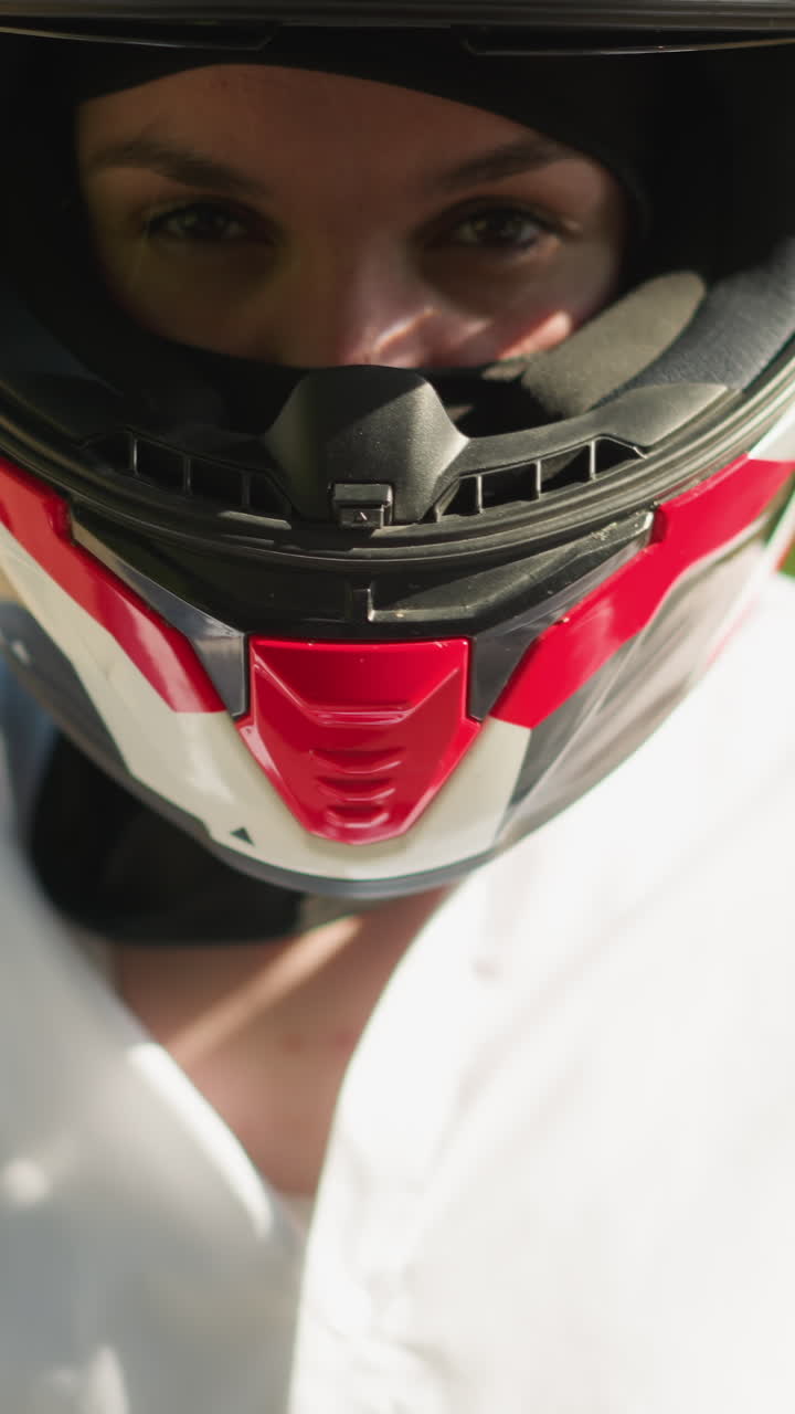 primer plano de una motociclista femenina con una camisa blanca y guantes negros ajustando y cerrando la visera de su casco, se aleja de la cámara con un fondo verde exuberante borroso