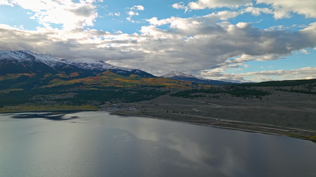 Aerial footage of a vast lake surrounded by green hills and snow-capped peaks in the distance