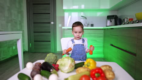 adorable toddler kid in jeans romper holding two big carrots. baby boy pointing at diverse vegetables learning them.