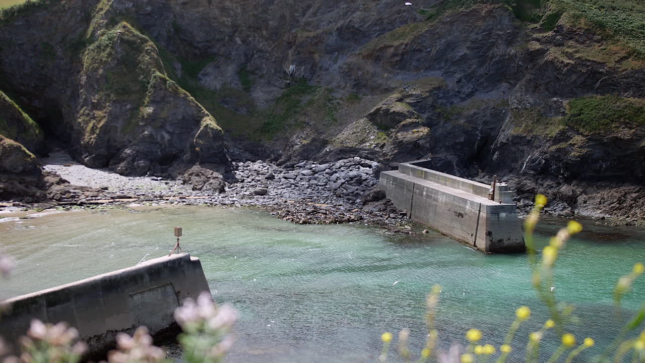 A harbour wall is stretching into clear shallow waters, backed by rugged cliffs in Cornwall near Port Isaac and the fictional village of Portwenn on the English coast