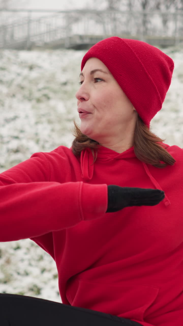 woman performs outdoor workout with hands up for balance turning left and right on snowy ground surrounded by snowy hill iron railing and blurred light pole under foggy atmosphere breath visible