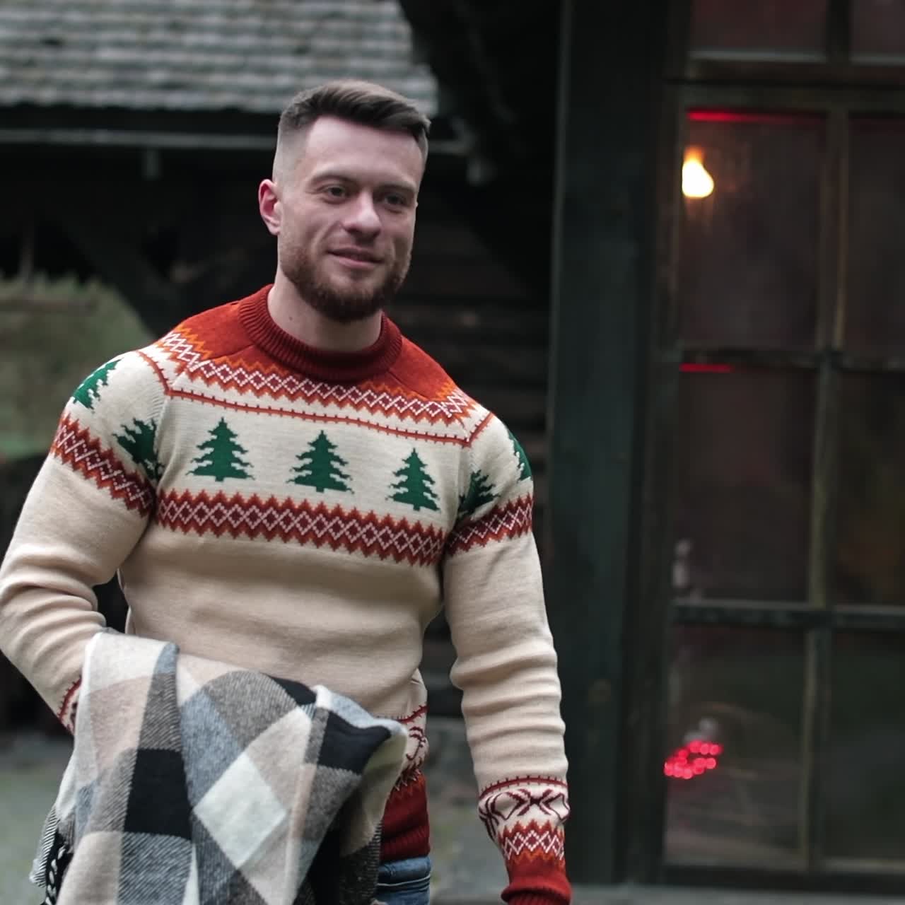 Smiling young man walks somewhere carrying warm blanket on his hand. Bearded male outside the big old rural house. Rural mansion with lights inside at the background