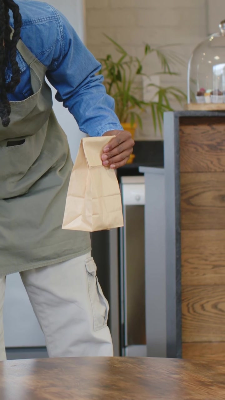 Barista che serve, indossa una camicia di jeans, grembiule verde oliva, appoggiato al bancone del caffè, tiene una tazza e un sacchetto di carta.