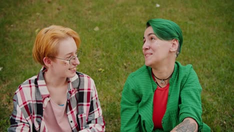 Close-up-shot-of-a-blonde-girl-with-a-short-haircut-with-glasses-in-a-checkered-pink-shirt-and-her-girlfriend-with-a-short-green-haircut-in-a-Green-shirt-and-a-red-T-shirt-are-sitting-on-the-lawn-on-green-grass-and-talking-about-something-during-their-meeting-in-the-park