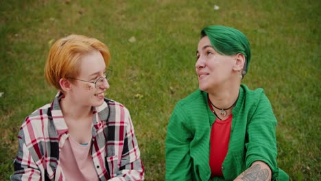 un primer plano de una chica rubia con un corte de pelo corto con gafas en una camisa rosada a cuadros y su novia con un corto corte de pelo verde en una camiseta verde y una camisa roja están sentados en el césped en la hierba verde y hablando de algo durante su reunión en el parque