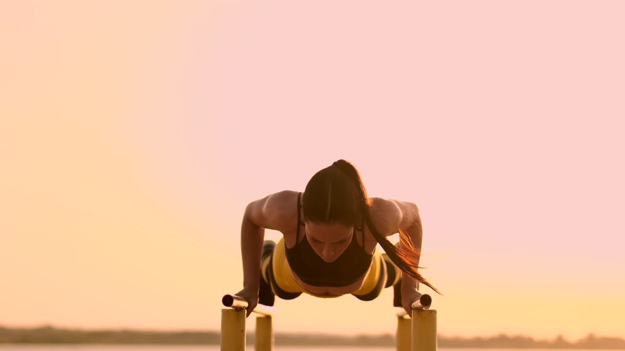 Beautiful slender woman athlete in a black top and yellow pants at sunset performs pushups on a parallel horizontal bar in slow motion.