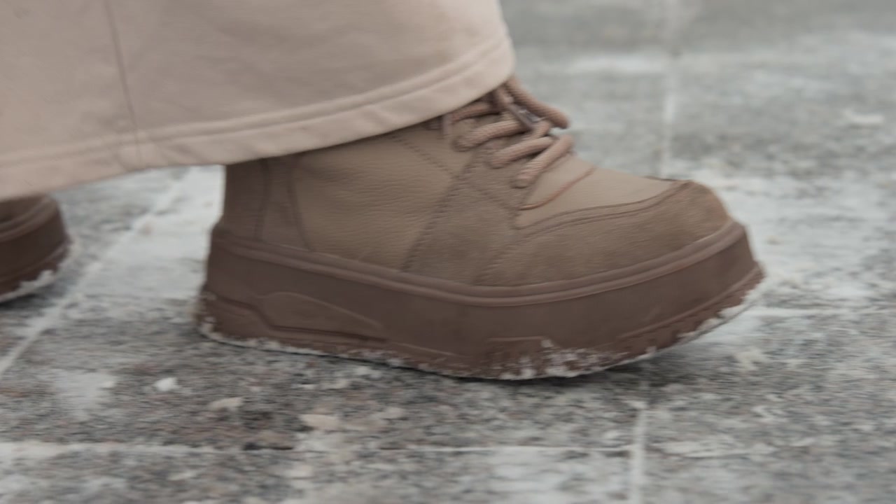 Low-angle close-up of two muddy brown boots walking on wet dark gray tile or stone, captured midstep with heel-to-toe roll, mud clinging to soles, and small water splashes along grout lines; diffuse