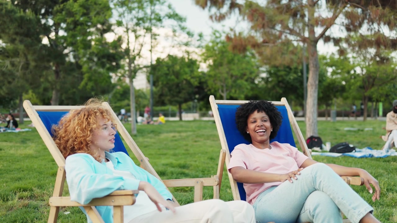 Two Diverse Young Women Relaxing and Talking in Park Deck Chairs