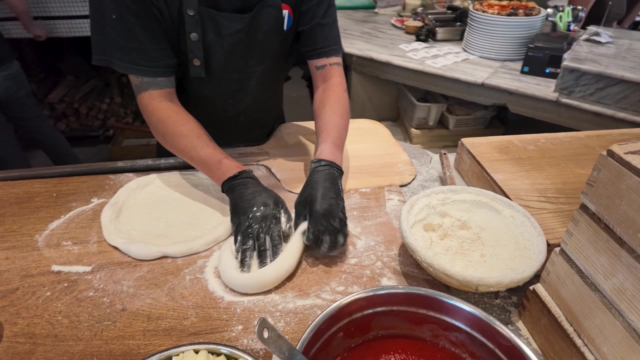 Chef usando luvas pretas prepara massa de pizza artesanal em superfície de madeira enfarinhada na cozinha do restaurante.