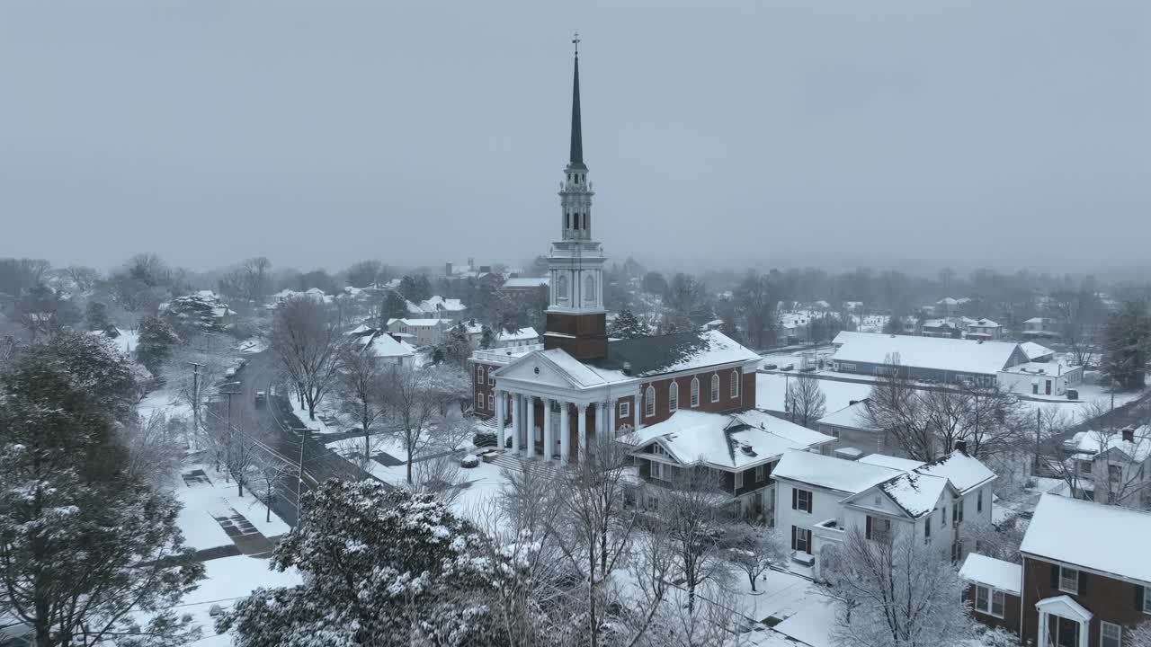 Sorvolo aereo di una città americana in inverno, che mostra case e alberi innevati con il campanile della chiesa che si erge sopra il quartiere sotto un cielo grigio e nuvoloso. Campo lungo