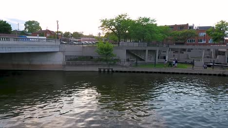 A-drone-flies-above-the-Flint-River-looking-toward-Riverbank-Park-in-Downtown-Flint,-Michigan-as-people-enjoy-a-summer-evening-at-dusk