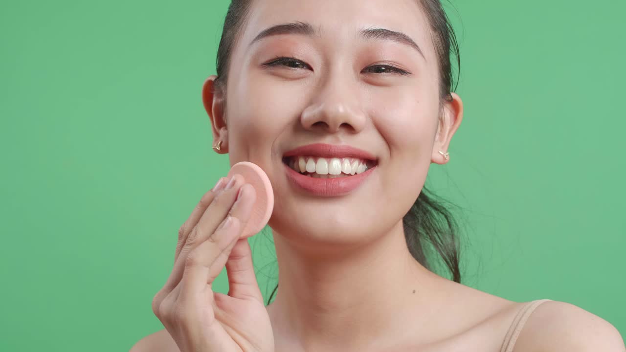 웰니스 스킨 케어를 위해 파우더 얼굴을 적용하고 스튜디오에서 녹색 화면 배경에 서있는 동안 미소 짓는 아시아 여성의 클로즈업